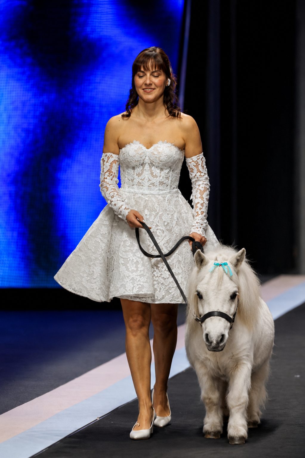 Modella in abito da sposa corto in pizzo sfila con un mini pony bianco